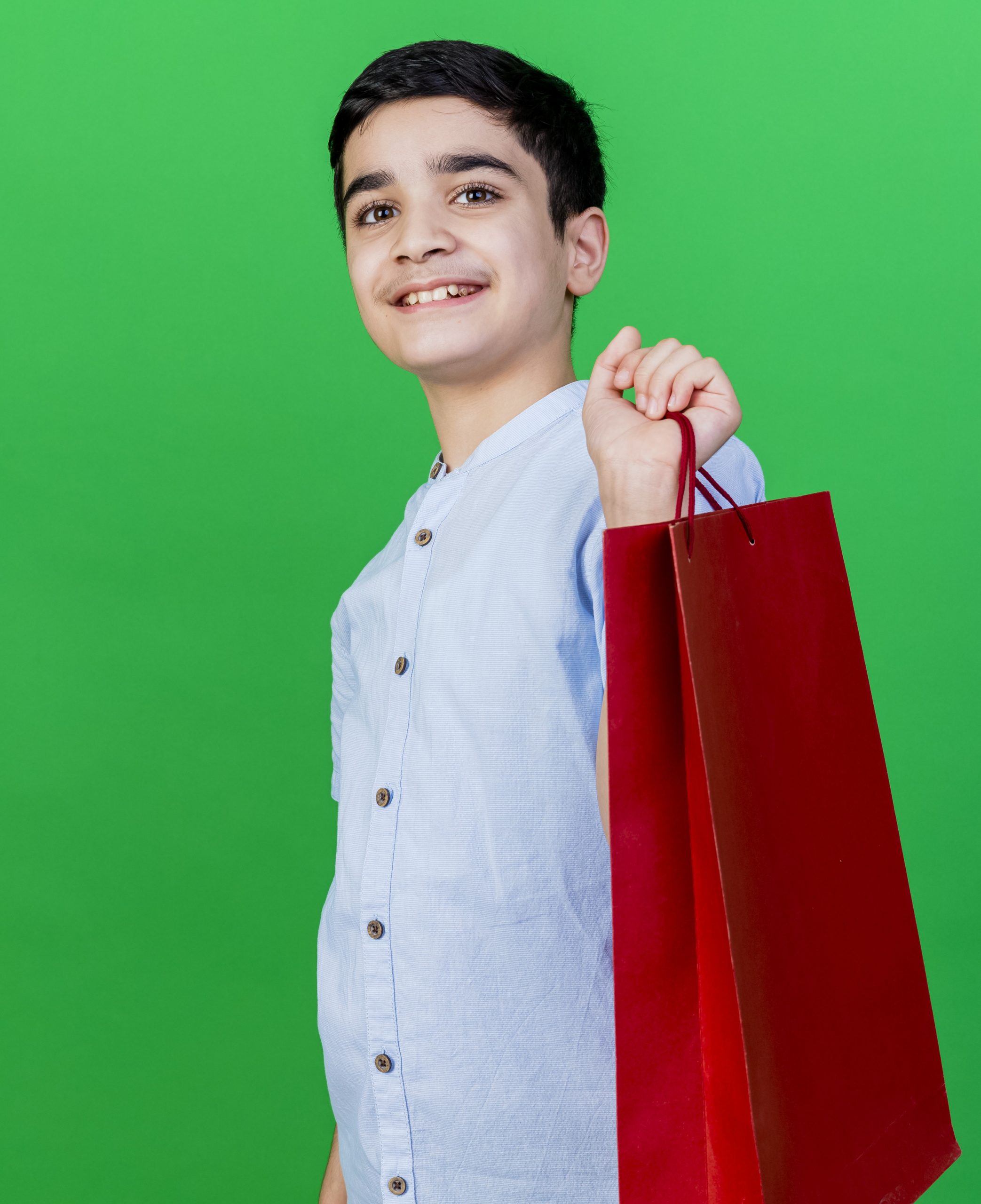 smiling young caucasian boy standing in profile view holding shopping bag looking at camera isolated on green background with copy space